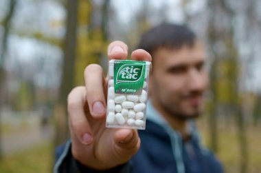 KHARKOV, UKRAINE - OCTOBER 26, 2019: Young man shows new Tic tac hard mints package in autumn park. Tic tac is popular due its minty fresh taste by Ferrero clipart