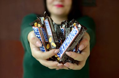 Kharkov, Ukrayna - 8 Ekim 2019: Kafkasyalı genç bir esmer kız karanlık odada birçok Snickers çikolatası gösteriyor. Mars yapımı Snickers çikolatası.