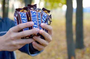 Kharkov, Ukrayna - 8 Ekim 2019: Genç bir beyaz adam sonbahar parkında kahverengi ambalajlı birçok Snickers çikolatası gösteriyor. Mars yapımı Snickers çikolatası.