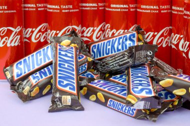 Kahverengi ambalajlı Snickers çikolataları parlak menekşe arkaplanda uzanır ve Coca Cola teneke kutuları kapanır. Meşhur içecek ve çikolata ürünü.