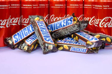 Kahverengi ambalajlı Snickers çikolataları parlak menekşe arkaplanda uzanır ve Coca Cola teneke kutuları kapanır. Meşhur içecek ve çikolata ürünü.