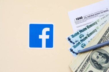 Facebook basılı logoda 1040 Bireysel Gelir Vergi İade Formu ve bej arka planda 100 Dolarlık banknotlar bulunuyor. Sosyal ağ kavramından vergi dönemine yardım