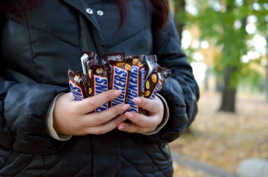 KHARKOV, UKRAINE - OCTOBER 8, 2019: A young caucasian woman shows many Snickers chocolate bars in brown wrapping in autumn park. Snickers chocolate manufactured by Mars
