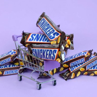 Birçok Snickers çikolatası açık mor arka planda alışveriş arabasıyla üst üste yığılmış. Snickers çikolataları Mars Anonim Şirketi tarafından üretilir.