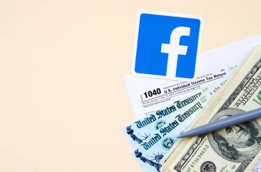 Facebook basılı logoda 1040 Bireysel Gelir Vergi İade Formu ve bej arka planda 100 Dolarlık banknotlar bulunuyor. Sosyal ağ kavramından vergi dönemine yardım