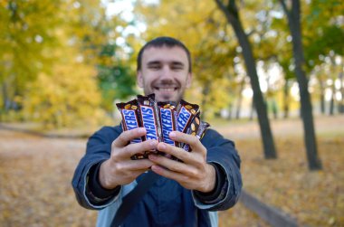 Kharkov, Ukrayna - 8 Ekim 2019: Beyaz sakallı genç bir adam sonbahar parkında kahverengi ambalajlı birçok Snickers çikolatası gösteriyor. Mars yapımı Snickers çikolatası.