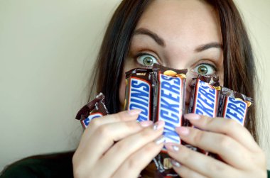 Kharkov, Ukrayna - 8 Ekim 2019: Genç, gülümseyen Kafkas esmer bir kız karanlık odada birçok Snickers çikolatası gösteriyor. Mars yapımı Snickers çikolatası.