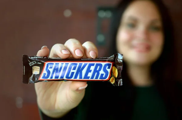 KHARKOV, UCRANIA - 8 de octubre de 2019: Una joven morena caucásica muestra una barra de chocolate Snickers en una habitación oscura. Snickers chocolate fabricado por Mars — Foto de Stock