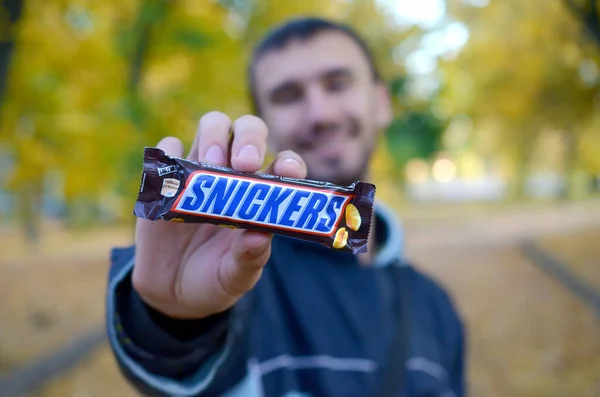 KHARKOV, UCRANIA - 8 DE OCTUBRE DE 2019: Un joven caucásico barbudo muestra una barra de chocolate Snickers en envoltura marrón en el parque de otoño. Snickers chocolate fabricado por Mars — Foto de Stock