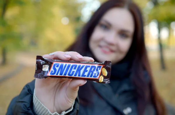 KHARKOV, UCRANIA - 8 de octubre de 2019: Una joven morena caucásica muestra una barra de chocolate Snickers en el parque de otoño. Snickers chocolate fabricado por Mars — Foto de Stock