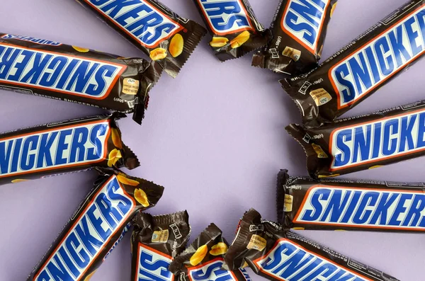 Muchas barras de chocolate Snickers se encuentra en papel pastel lila. Las barras Snickers son producidas por Mars Incorporated. Snickers fue creado por Franklin Clarence Mars en 1930 — Foto de Stock