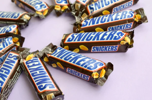 Muchas barras de chocolate Snickers se encuentra en papel pastel lila. Las barras Snickers son producidas por Mars Incorporated. Snickers fue creado por Franklin Clarence Mars en 1930 — Foto de Stock