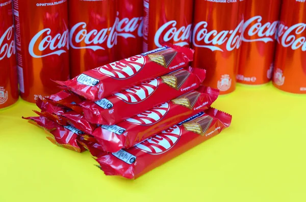 Kit Kat barras de chocolate en envoltura roja se encuentra sobre fondo amarillo brillante con latas de Coca Cola de cerca. Famoso producto de bebida y chocolate —  Fotos de Stock