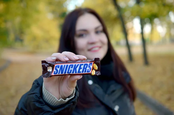 KHARKOV, UCRANIA - 8 de octubre de 2019: Una joven morena caucásica muestra una barra de chocolate Snickers en el parque de otoño. Snickers chocolate fabricado por Mars —  Fotos de Stock