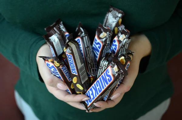 KHARKOV, UCRÂNIA - OUTUBRO 8, 2019: Uma jovem morena caucasiana mostra muitas barras de chocolate Snickers no quarto escuro. Chocolate Snickers fabricado pela Mars — Fotografia de Stock