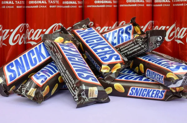Snickers chocoladerepen in bruine omwikkeling liggen op heldere violette achtergrond met Coca Cola blikjes dicht. Bekende drank en chocoladeproduct — Stockfoto
