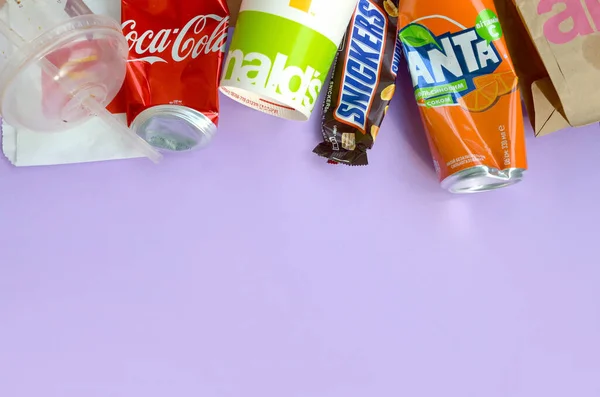 Snickers chocolade bar verpakking ligt met lege rode Coca Cola en oranje Fanta blikken naast mcdonalds papieren zakken en kopjes op lila pastel achtergrond. Beroemde drank en chocolade product afval — Stockfoto