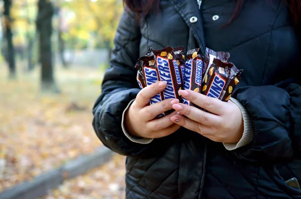 KHARKOV, UCRANIA - 8 DE OCTUBRE DE 2019: Una joven caucásica muestra muchas barras de chocolate Snickers en envoltura marrón en el parque de otoño. Snickers chocolate fabricado por Mars — Foto de Stock