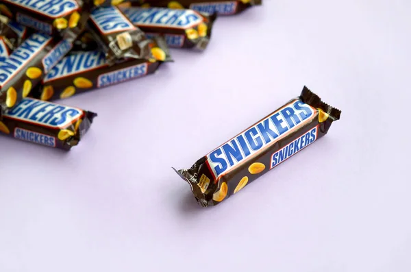 Muchas barras de chocolate Snickers se encuentra en papel pastel lila. Las barras Snickers son producidas por Mars Incorporated. Snickers fue creado por Franklin Clarence Mars en 1930 — Foto de Stock