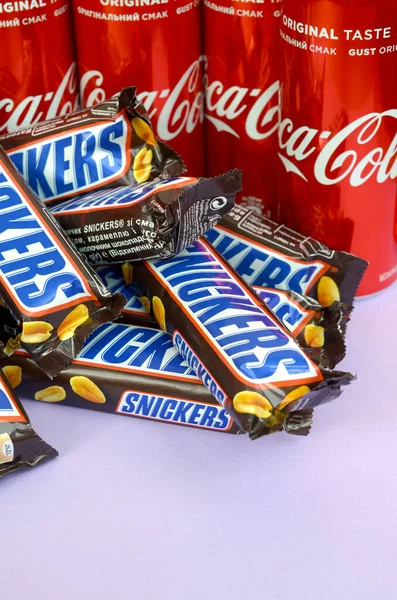 Snickers čokoládové tyčinky v hnědém obalu leží na jasně fialovém pozadí s Coca Cola plechovky zblízka. Slavný nápoj a čokoládový výrobek — Stock fotografie
