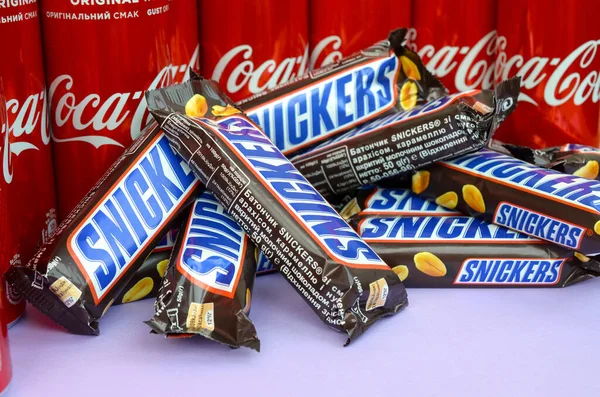 Snickers sjokolade i brun innpakning ligger på lys fiolett bakgrunn med Coca Cola blikkbokser som lukker seg. Berømte drikkevarer og sjokoladeprodukter – stockfoto