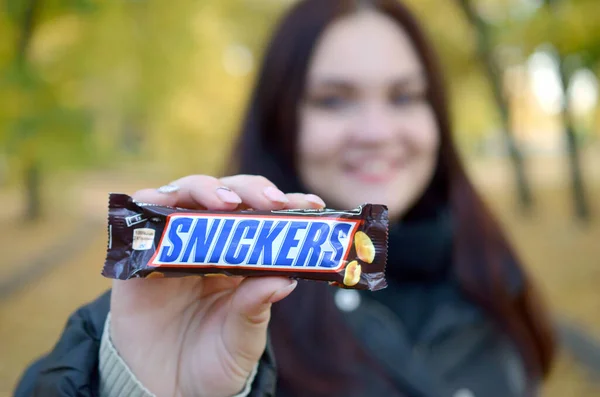 KHARKOV, UCRANIA - 8 de octubre de 2019: Una joven morena caucásica muestra una barra de chocolate Snickers en el parque de otoño. Snickers chocolate fabricado por Mars — Foto de Stock