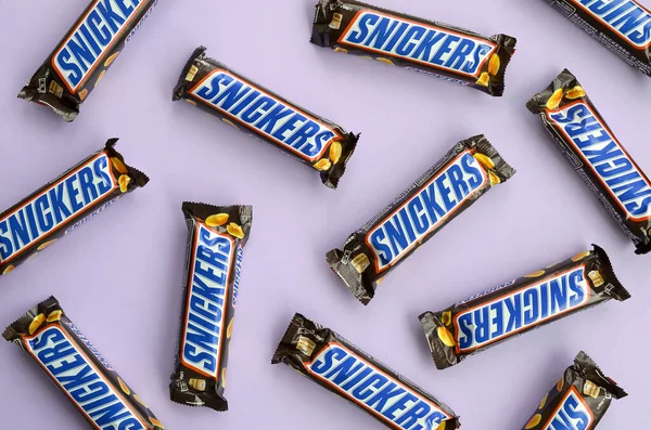 Mnoho čokoládových tyčinek Snickers leží na pastelovém šeříkovém papíru. Tyčinky Snickers vyrábí společnost Mars Incorporated. Snickers vytvořil Franklin Clarence Mars v roce 1930 — Stock fotografie
