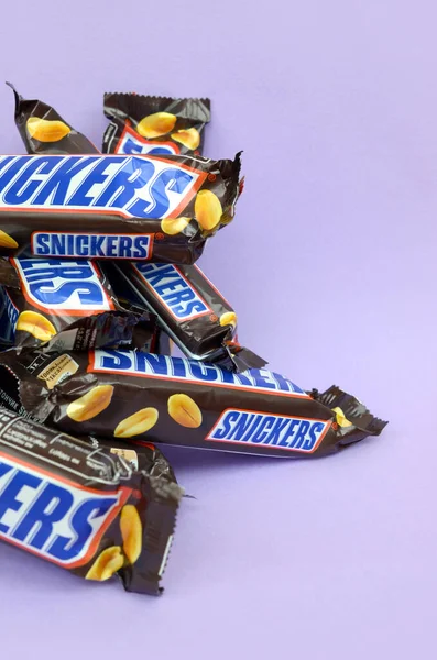 Многие шоколадные батончики Snickers лежат на пастельной лиловой бумаге. Сникерс-бары производятся Mars Incorporated. Snickers был создан Франклином Кларенсом Марсом в 1930 году. — стоковое фото