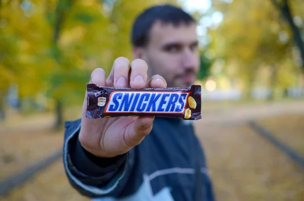 KHARKOV, UCRANIA - 8 DE OCTUBRE DE 2019: Un joven caucásico barbudo muestra una barra de chocolate Snickers en envoltura marrón en el parque de otoño. Snickers chocolate fabricado por Mars — Foto de Stock