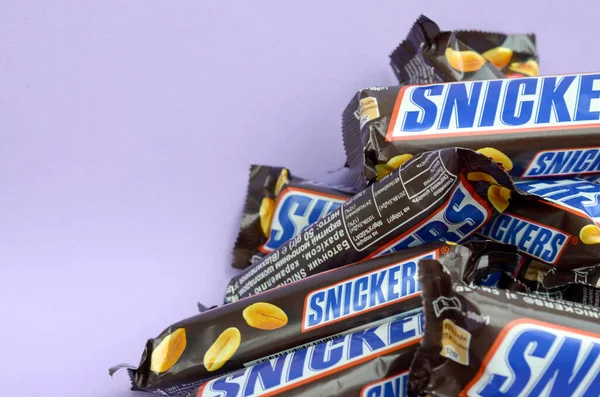 Muchas barras de chocolate Snickers se encuentra en papel pastel lila. Las barras Snickers son producidas por Mars Incorporated. Snickers fue creado por Franklin Clarence Mars en 1930 — Foto de Stock