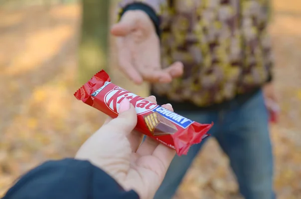 KHARKOV, UCRÂNIA - OUTUBRO 21, 2019: Mão masculina passa a menina uma barra de chocolate Kit kat em um parque de outono. A manifestação da bondade, tratando com doces — Fotografia de Stock