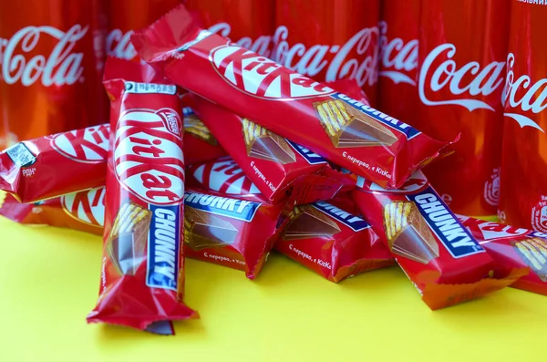 Kit Kat barras de chocolate em vermelho embrulho encontra-se no fundo amarelo brilhante com latas de lata Coca Cola de perto. Bebida famosa e produto de chocolate — Fotografia de Stock