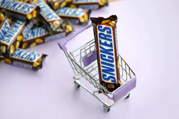 Muitas barras de chocolate Snickers empilhadas de perto com carrinho de compras em fundo violeta claro. Barras Snickers são produzidas pela Mars Incorporated — Fotografia de Stock