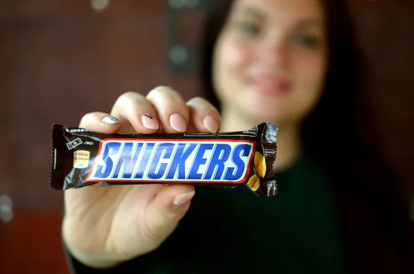KHARKOV, UCRANIA - 8 de octubre de 2019: Una joven morena caucásica muestra una barra de chocolate Snickers en una habitación oscura. Snickers chocolate fabricado por Mars —  Fotos de Stock