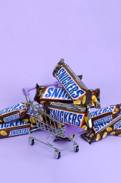 Många Snickers chokladkakor staplade på nära håll med kundvagn på ljus violett bakgrund. Snickers barer tillverkas av Mars Incorporated — Stockfoto