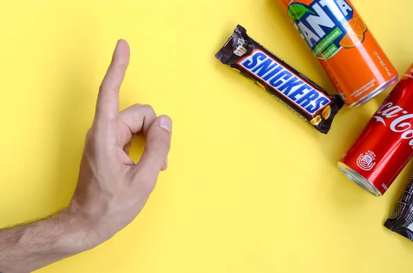 Puntas de mano masculinas en barra de chocolate Snickers en envoltura marrón, lata de Coca Cola roja y Fanta naranja sobre fondo pastel amarillo. Famoso producto de bebida y chocolate —  Fotos de Stock