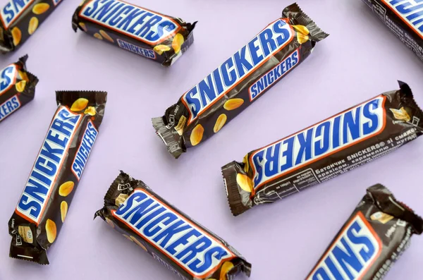 Muchas barras de chocolate Snickers se encuentra en papel pastel lila. Las barras Snickers son producidas por Mars Incorporated. Snickers fue creado por Franklin Clarence Mars en 1930 — Foto de Stock