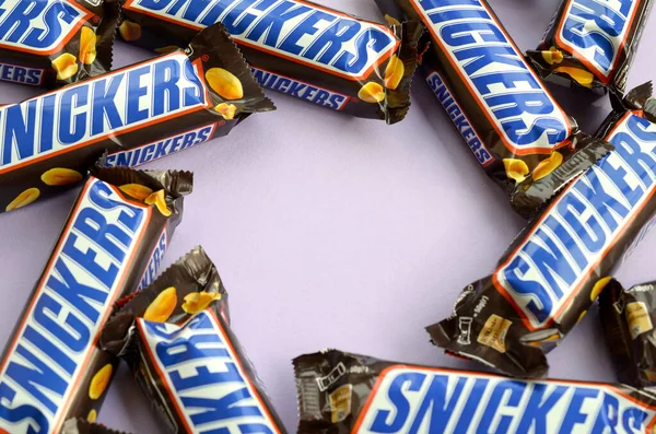 Mnoho čokoládových tyčinek Snickers leží na pastelovém šeříkovém papíru. Tyčinky Snickers vyrábí společnost Mars Incorporated. Snickers vytvořil Franklin Clarence Mars v roce 1930 — Stock fotografie