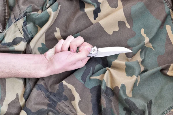 迷彩生地の背景に小さなナイフで手。兵士の間で近接戦術にナイフを使用するという概念 — ストック写真