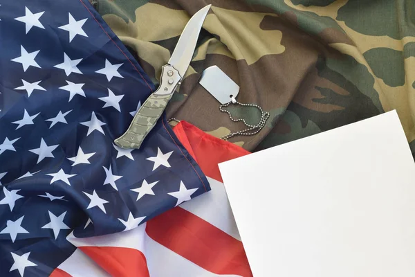 Blank paper lies with knife and army dog tag necklace on camouflage uniform and american flag — Stock Photo, Image