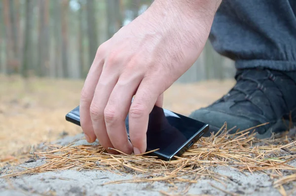 Male hand picking up lost mobile phone from a ground in autumn fir wood path. The concept of finding a valuable thing and good luck