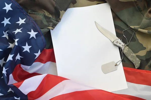 Blank paper lies with knife and army dog tag necklace on camouflage uniform and american flag — Stock Photo, Image