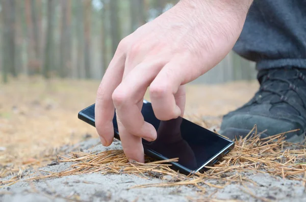 Male hand picking up lost mobile phone from a ground in autumn fir wood path. The concept of finding a valuable thing and good luck