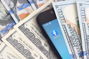 Kharkov, Ukrayna - 6 Şubat 2020: Facebook uygulamalı akıllı telefon ekranı ve bir sürü yüz dolarlık banknot. İş ve sosyal ağ kavramı