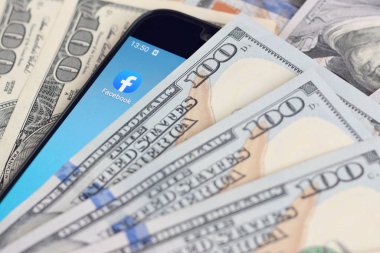 Kharkov, Ukrayna - 6 Şubat 2020: Facebook uygulamalı akıllı telefon ekranı ve bir sürü yüz dolarlık banknot. İş ve sosyal ağ kavramı