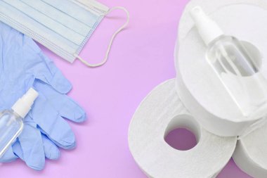 Covid-19 karantina saatleri için önemli eşyalar. Tuvalet kağıdı, plastik tek kullanımlık eldivenler, ameliyat maskesi ve eflatun arka planda el dezenfektanı.