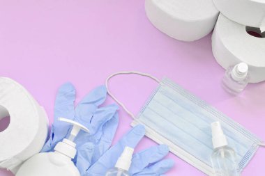 Covid-19 karantina saatleri için önemli eşyalar. Tuvalet kağıdı, ameliyat maskeli tek kullanımlık eldivenler ve eflatun arkaplanda sıvı sabun şişesi olan el dezenfektanı.