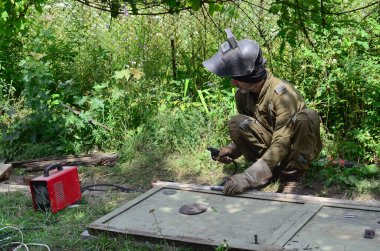 Kahverengi üniformalı yaşlı kaynakçı, kaynak maskesi ve kaynakçı derisi, kaynak makinesi olan metal kapılar açık havada, mavi kıvılcımlar yanlarda uçuşuyor.