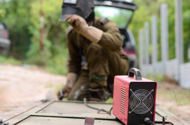 Kahverengi üniformalı yaşlı kaynakçı, kaynak maskesi ve kaynakçı derisi, kaynak makinesi olan metal kapılar açık havada, mavi kıvılcımlar yanlarda uçuşuyor.