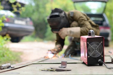 Kahverengi üniformalı yaşlı kaynakçı, kaynak maskesi ve kaynakçı derisi, kaynak makinesi olan metal kapılar açık havada, mavi kıvılcımlar yanlarda uçuşuyor.